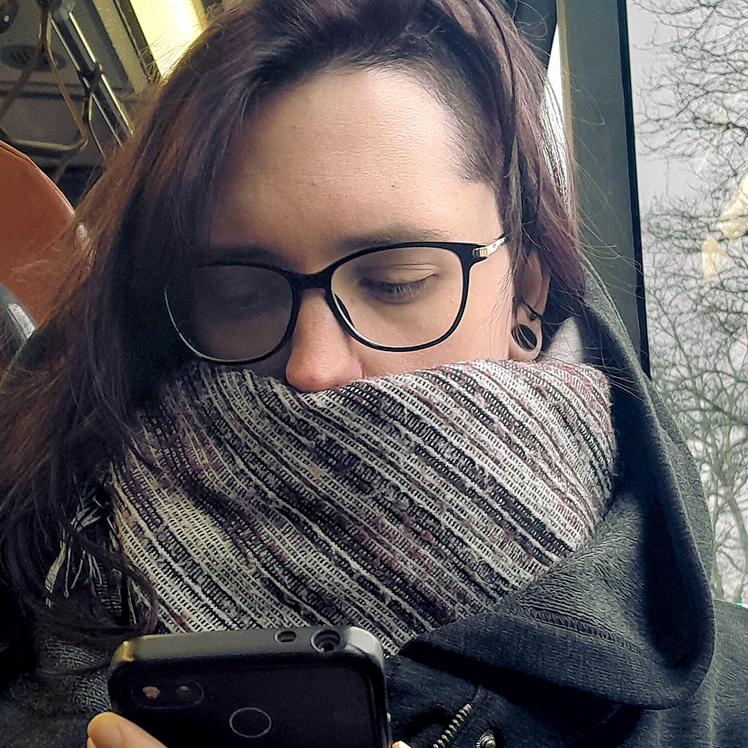 Photo dans un bus, regardant son téléphone. Cheveux un peu rouge/bordeaux avec des lunettes noires. On entrevoit un tunnel lobe gauche noir ainsi qu&rsquo;un conche.